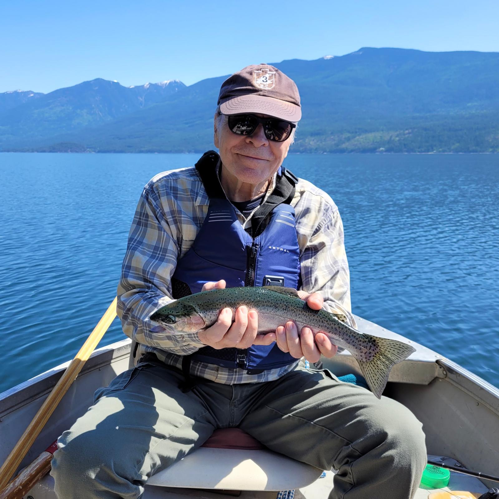 James Utley holding a rainbow trout he has caught, seated in a boat on a lake with forested mountains behind him